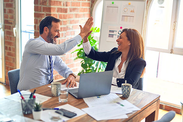 Ein Mann und eine Frau sitzen an einem Schreibtisch in einem modernen Büro mit Ziegelwand und Fenstern. Beide lachen und geben sich ein High-Five. Auf dem Tisch liegen ein Laptop, Dokumente, Tassen und Bürozubehör. Im Hintergrund sind Pflanzen und ein Flipchart mit Diagrammen zu sehen.
