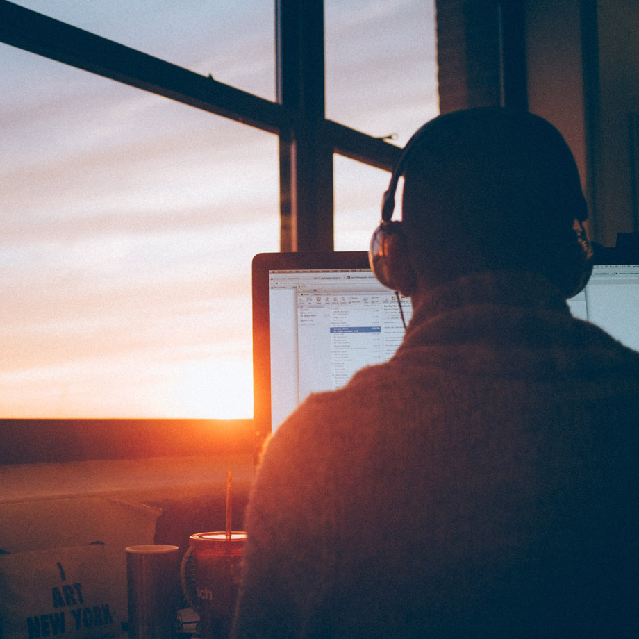 Eine Person mit Kopfhörern sitzt an einem Schreibtisch vor einem Computerbildschirm, auf dem ein E-Mail-Programm geöffnet ist. Im Hintergrund scheint die Sonne durch ein großes Fenster.