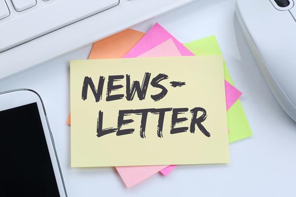 Ein gelber Notizzettel mit der handgeschriebenen Aufschrift "NEWSLETTER" liegt auf bunten Haftnotizen neben einer Computermaus und einem Smartphone.